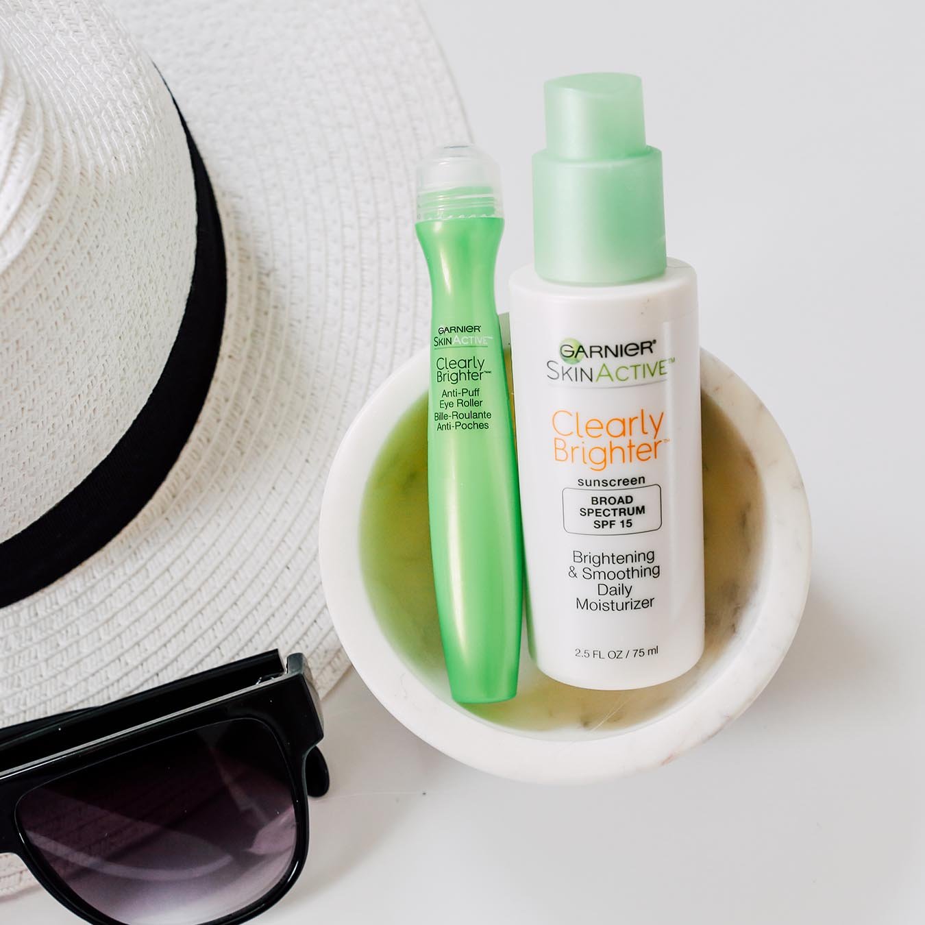 Garnier SkinActive Clearly Brighter Anti-Puff Eye Roller and SkinActive Clearly Brighter Sunscreen Broad Spectrum SPF 15 Brightening & Smoothing Daily Moisturizer in a white marble bowl on a white background next to a white straw hat with black band and black sunglasses.